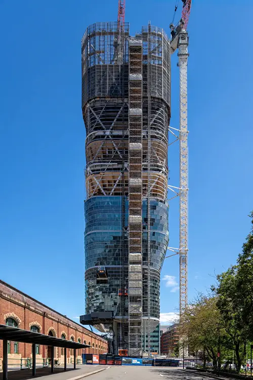 Ein moderner Wolkenkratzer im Bau, umgeben von Baugerüsten und einem Kran oben, mit reflektierenden Glasfassaden vor blauem Himmel.