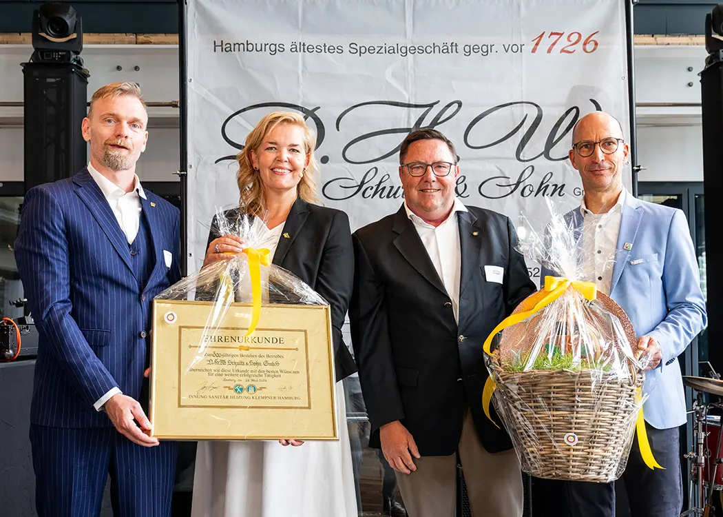 Zwei Personen in formeller Kleidung stehen nebeneinander vor einem Schild, das teilweise im Hintergrund sichtbar ist.