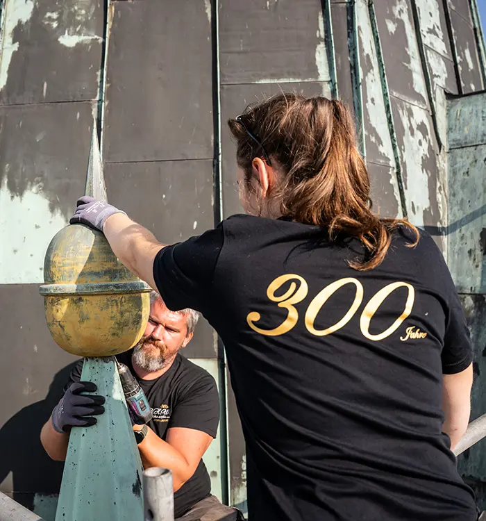 Eine Person mit dunklem Haar trägt ein schwarzes T-Shirt mit gelber Aufschrift, steht im Freien vor einer Wand, teilweise von hinten zu sehen.