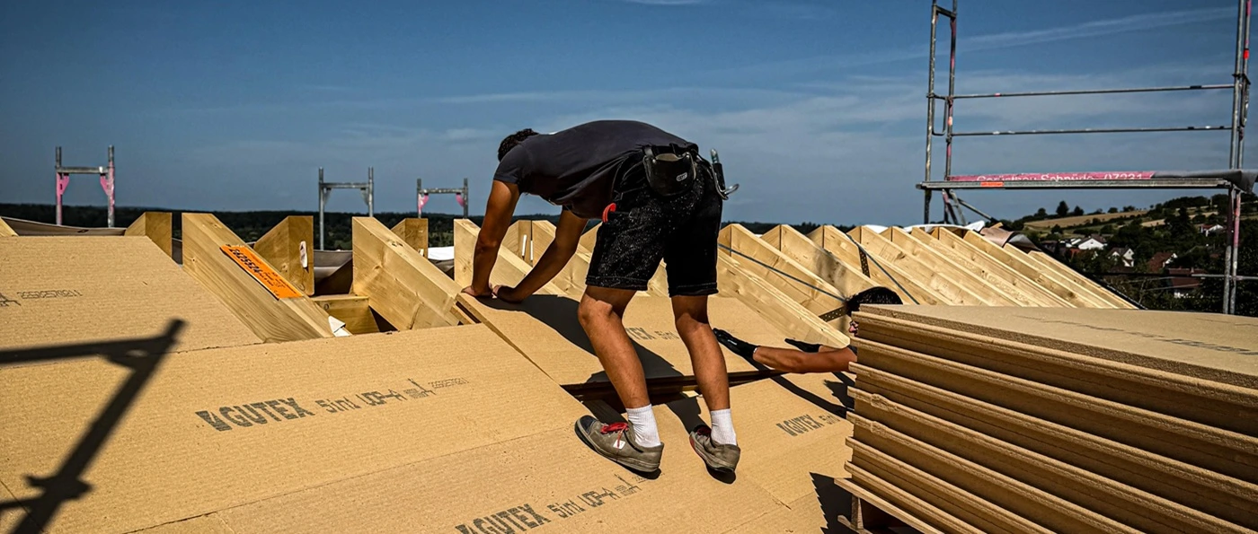 Dettinger-Titelbild Ein Arbeiter in Shorts und Sportschuhen kniet auf einem Dach mit Holzplatten unter blauem Himmel.