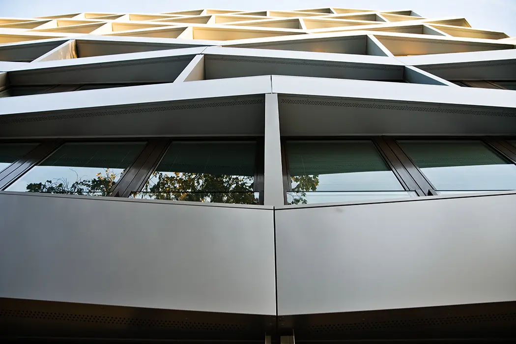Moderne Gebäudefassade mit Glas- und Metallelementen, reflektierende Fenster, strukturierte Linien und etwas Grün im Hintergrund.