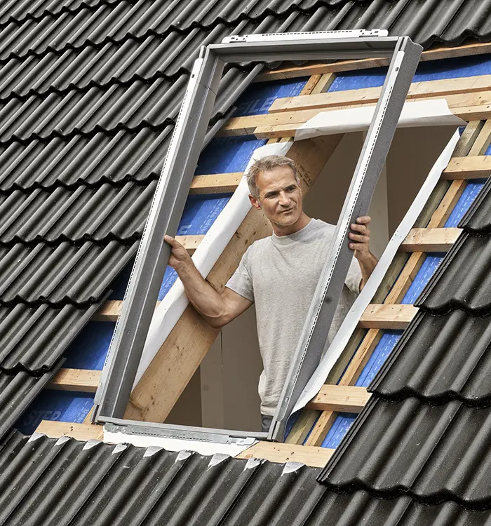 Ein Mann steht auf einer Leiter und arbeitet am Dach eines Hauses, während er Baumaterialien hält und trägt.