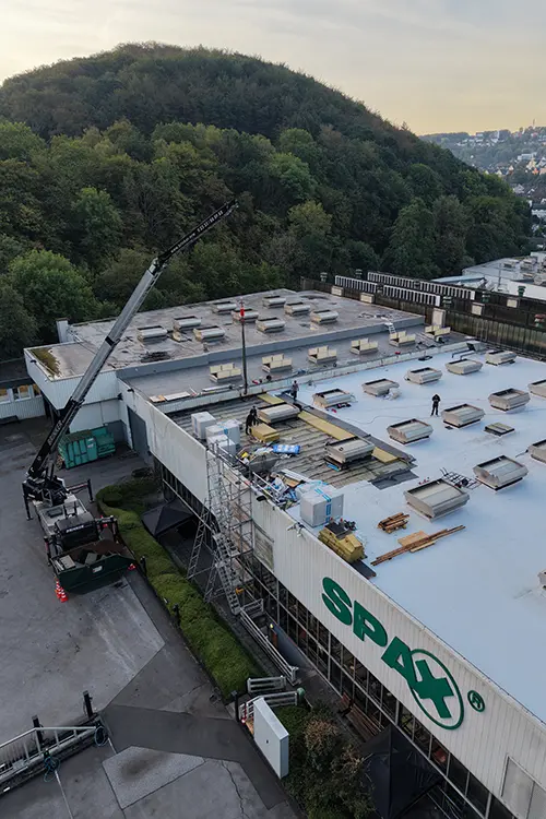 Arbeiter renovieren das Dach eines Industriegebäudes mit Baumaterialien, Gerüst und Kran, umgeben von grünen Bäumen und Hügeln im Hintergrund.