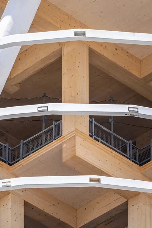 Holzkonstruktion mit mehreren Etagen, rechte Winkel und Balkone, Metallgeländer und sichtbaren Verbindungselementen, moderne Architektur und helle Holzoberflächen.