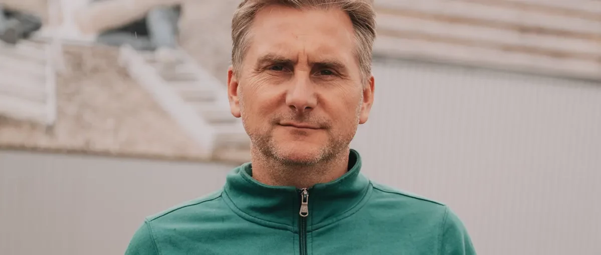 Nahaufnahme eines Mannes mit kurzen Haaren, leichtem Bart und grünem Reißverschlussjacke, neutraler Gesichtsausdruck, Hintergrund unscharf.