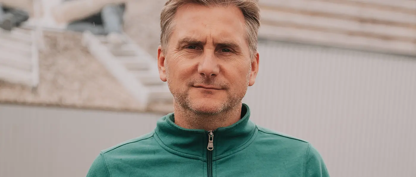 Nahaufnahme eines Mannes mit kurzen Haaren, leichtem Bart und grünem Reißverschlussjacke, neutraler Gesichtsausdruck, Hintergrund unscharf.