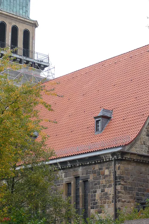 Steingebäude mit großen Fenstern und einem roten Ziegeldach, teilweise von Bäumen verdeckt, bei Tageslicht aufgenommen.