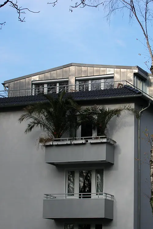 Modernes Wohngebäude mit großen Fenstern und Balkon, teilweise von Palmenpflanzen verdeckt, unter blauem Himmel.
