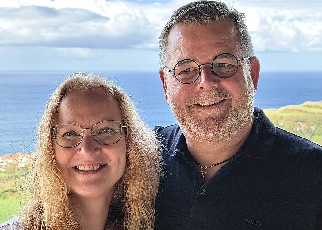 Ein Mann mit Brille und Bart steht draußen, im Hintergrund ist das Meer und blauer Himmel mit Wolken zu sehen.