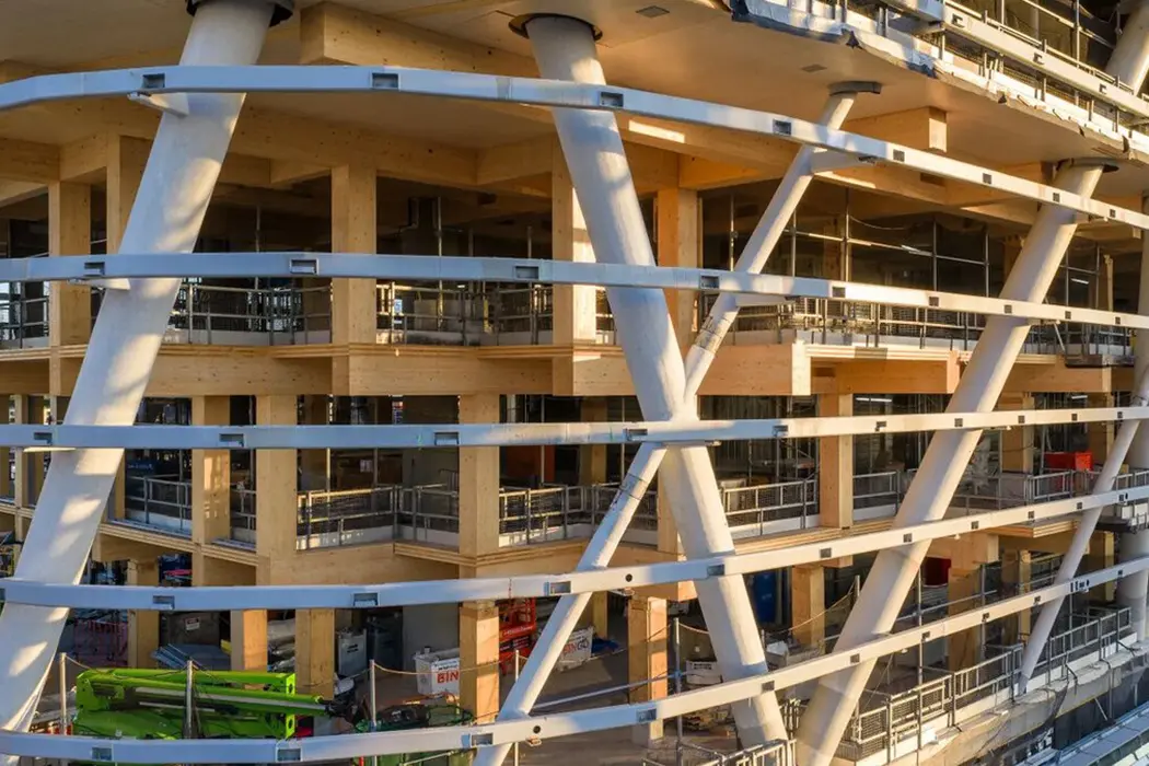 Mehrstöckiges Gebäude im Bau mit sichtbaren Stahlträgern und Holzstrukturen, teilweise eingerüstet, Sonnenlicht fällt auf die Baustelle.