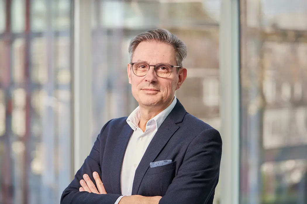 Ein Mann mit Brille, Anzug und weißem Hemd blickt freundlich in die Kamera, Hintergrund unscharf.