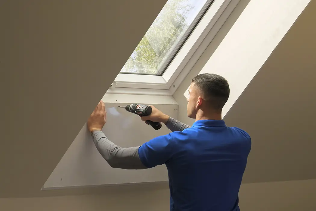 Eine Person in blauer Kleidung benutzt einen Akkuschrauber und arbeitet an einem Fenster in einem hellen Raum.