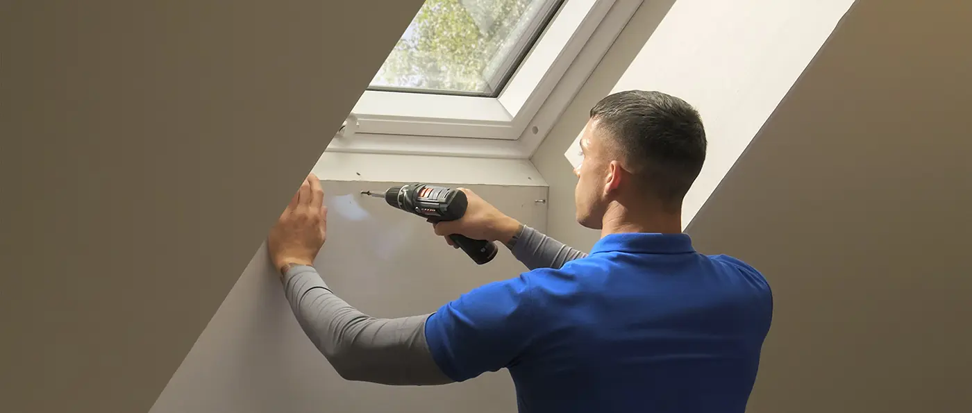 Eine Person hält eine elektrische Bohrmaschine und arbeitet an einer Wand unter einem Dachfenster.