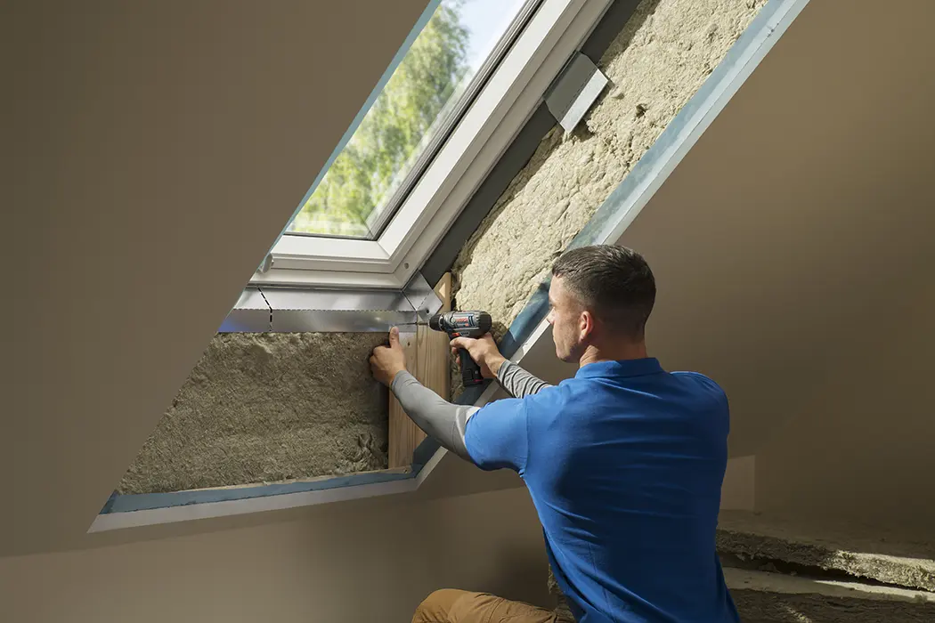 Person verwendet eine Bohrmaschine, um ein Holzbrett an einer Dachschräge nahe einem Fenster zu befestigen. Tageslicht scheint herein.